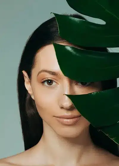 Frau schaut durch ein großes grünes Blatt, symbolisiert natürliche Schönheit und Pflege